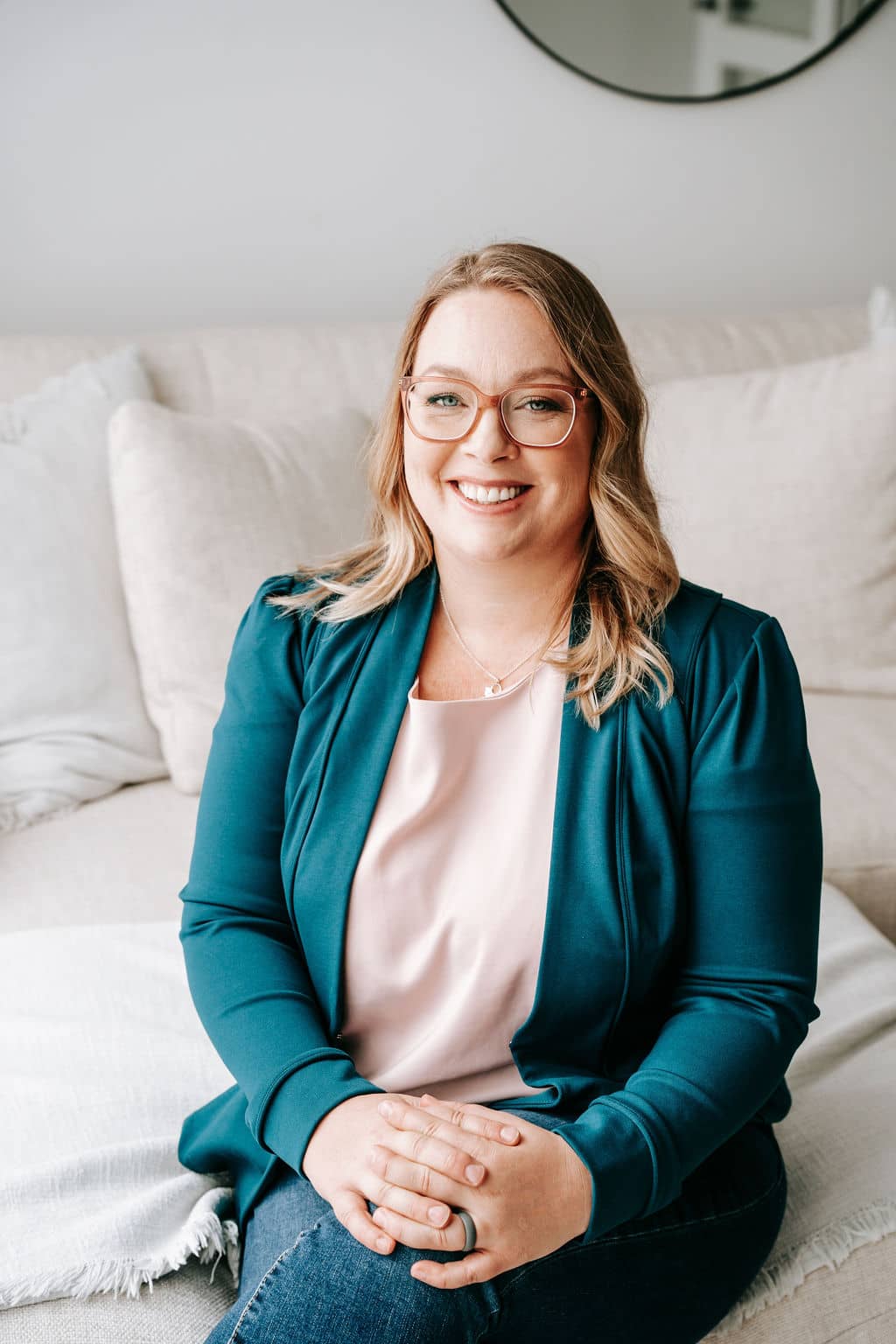 Kate, the owner of Kip & Boo, sitting cross-legged on the edge of a couch, smiling at the camera with glasses on, embodying the brand's welcoming and authentic spirit