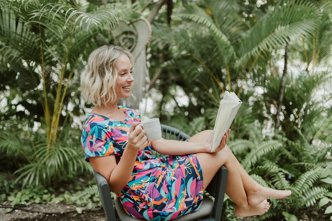 Women sitting sideways on chair with coffee in hand and reading a book in kip and boo bamboo nightie in cancun print
