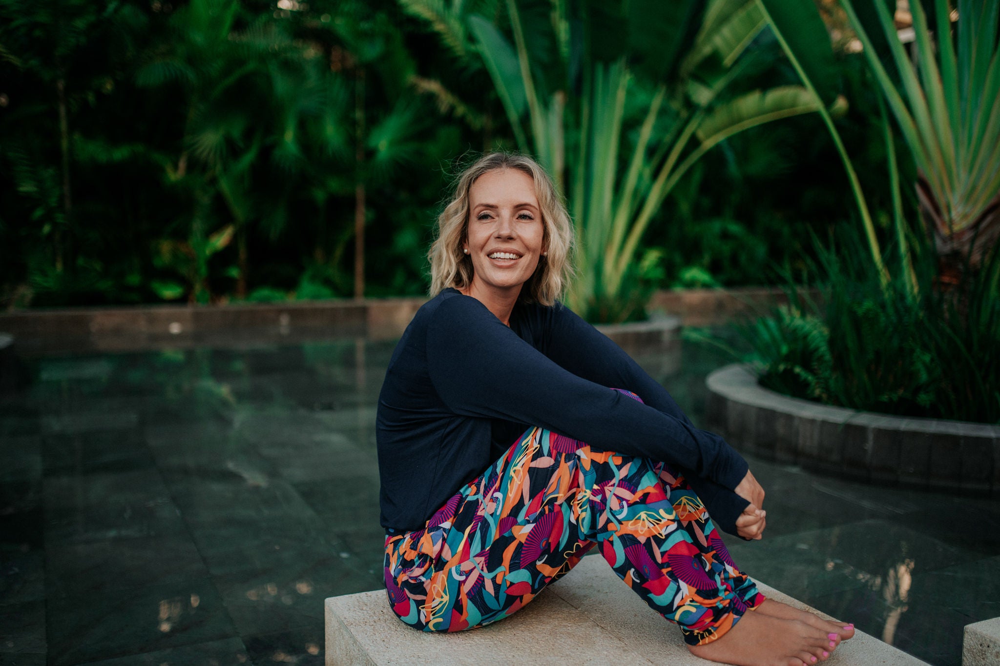 Model in Kip & Boo's Cancun bamboo pants and Seminyak top sitting on a ledge over a pool with lush tropical palm trees and garden in the background, showcasing eco-friendly and stylish sleepwear.